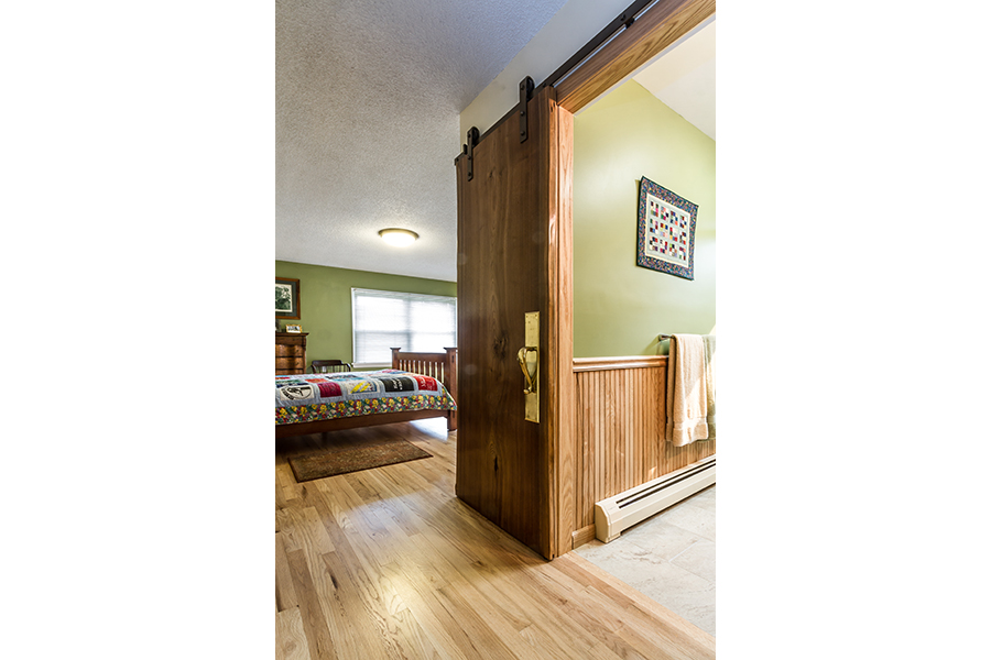 A sliding wooden barn-door style entrance leads to a bathroom from a bedroom with no threshold. The bedroom has light wood floors and a quilted bed, while the bathroom has a linoleum floor and towels hanging above a radiator on the floor.